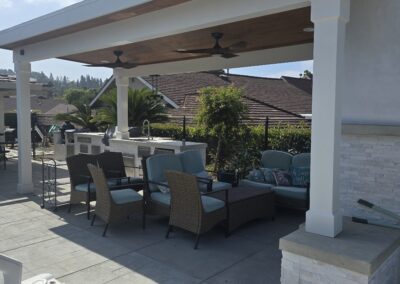 Covered patio with outdoor seating and a ceiling fan beside a pool, with a built-in grill and greenery visible in the background.
