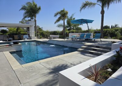A modern backyard features a rectangular swimming pool with water jets, lounge chairs under a blue umbrella, palm trees, and a covered patio area.